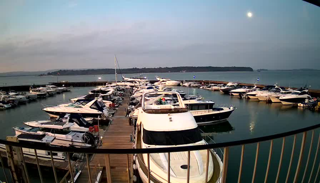 A marina with numerous boats docked at their slips, some covered with navy blue tarps. The water is calm and reflective, mirroring the boats and the pale blue sky above. In the background, a shoreline with greenery is visible. A full moon is positioned in the sky, casting a soft light over the scene.