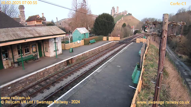 A quiet railway station platform is shown under early morning light. The platform features stone walls and a peaked roof with a chimney. There are several green benches and a green shed in the background. A sign reading "WAY OUT" is visible. In the distance, a hill with castle ruins can be seen. The railway tracks are in the foreground, running parallel to the platform. A wooden pole with wires stands to the right of the image. The scene conveys a peaceful, rural setting.