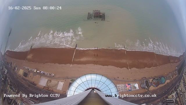 Aerial view looking down from a high vantage point. The image shows a sandy beach with waves gently lapping at the shore. There is a pier with some structures partially submerged in water. The beach is lined with several beach huts and an amusement area on the left, featuring colorful patterns. The coastline extends further along, with a road visible behind the beach, and cloudy skies above. The timestamp indicates it is Sunday, February 16, 2025, at 8:00 AM.