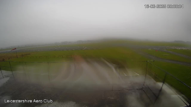 A blurry image of an airfield under cloudy and foggy weather conditions. The landscape is predominantly green grass, with a section of the airstrip visible in the distance. There's a partial view of a red flag on a pole, indicating wind direction, and the text "Leicestershire Aero Club" appears at the bottom. Rain or moisture is affecting the camera lens, causing a hazy view. The date and time displayed on the top right corner reads "16-02-2025 08:00:04."