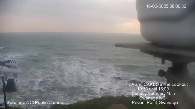 A coastal view showing turbulent waves crashing against rocky cliffs. The sky is overcast with dull gray clouds, suggesting a gloomy day. In the foreground, a metal structure is partially visible on the right, with directional markers marked with black and white stripes. The lower part of the image includes some grass and ground, while the water is a mix of dark blue and gray. The bottom corner features text promoting a tea and cakes event, along with details about its timing and location in Swanage.