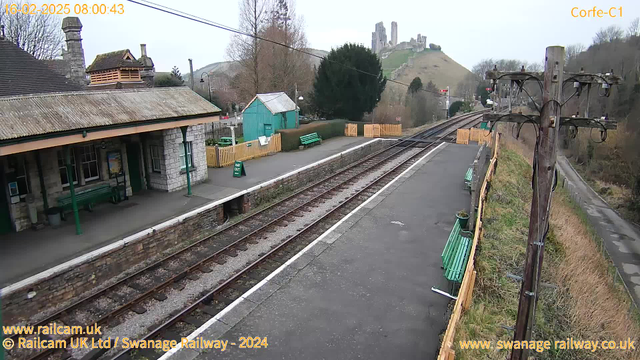 A railway station platform is visible with a stone building featuring a sloped roof on the left. There are several green benches along the platform and a wooden fence in the background. A small blue structure is nearby, and a green sign reading "WAY OUT" is positioned on the ground. In the background, a hill rises, with the ruins of a castle on top. There are railway tracks leading off to the right, with a wooden telephone pole and overhead wires nearby. The sky is overcast.