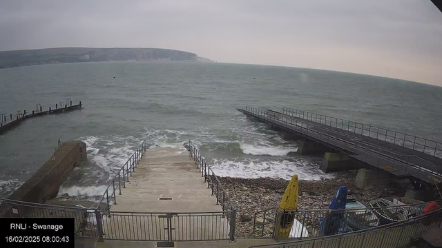 A view from a webcam showing a coastal scene. In the foreground, there is a set of concrete steps leading down to the water, bordered by a metal railing. To the right, a wooden pier extends over the waves, with some boats visible at the end. The water appears choppy and blue, with white foam where the waves break. In the center of the scene, there are pebbles along the shore. Two kayaks, one yellow and one blue, are positioned on the right side, next to a wall. The sky is overcast and gray, indicating a cloudy day.