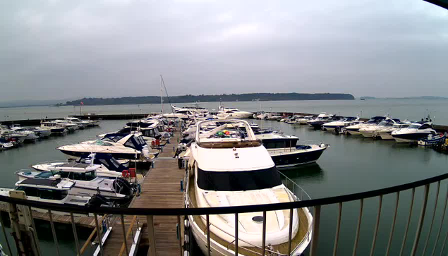 A marina scene featuring numerous boats docked in calm water. The foreground shows a wooden dock with a railing, while various boats of different sizes are moored in the harbor. The sky is overcast, and there is a faint outline of land in the background. The overall atmosphere appears tranquil and slightly misty.