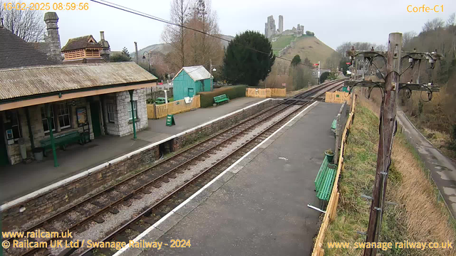 A view of Corfe Castle train station, featuring a stone platform with several green benches. The platform has a wooden fence on one side and a sign indicating "WAY OUT." In the background, there are rolling green hills, and the ruins of Corfe Castle are visible on the hilltop. It's a cloudy day, and train tracks run alongside the platform, leading off into the distance. Power lines are present on the right side of the image.