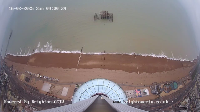 Aerial view of a beach with brown sand and gentle waves lapping at the shore. In the center, a partially submerged old pier structure is visible. To the right, colorful beach attractions and a pool can be seen, along with a road lined by buildings. The image has timestamps and text indicating it is powered by Brighton CCTV.