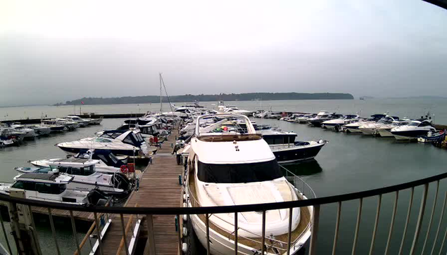 A view of a marina filled with various boats and yachts docked along wooden piers. The water is calm, reflecting the boats. The sky is overcast, giving a grayish tone to the scene. In the background, there are green hills visible on the horizon. A few flags are seen, possibly on the docks or boats. The overall atmosphere appears calm and peaceful.