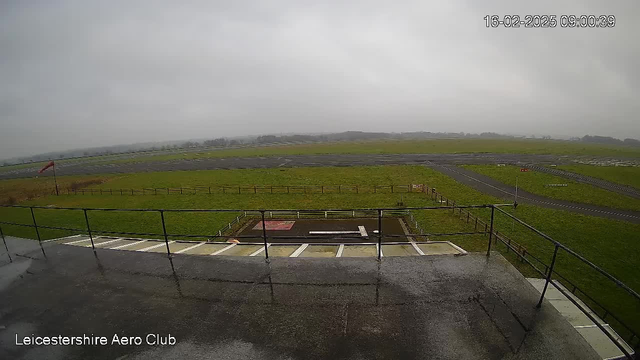 A view from a webcam at Leicestershire Aero Club shows an expansive green field under a gray overcast sky. The image captures part of the club's building with a railing in the foreground, and a wet surface indicates recent rain. In the background, a runway is visible, along with a red windsock indicating wind direction. The area is mostly empty, with a few scattered fences around the grassy landscape.