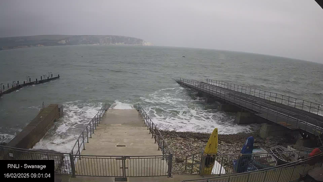 A cloudy coastal scene showing rough waves crashing against a rocky shoreline. In the foreground, there are concrete steps leading down toward the water, flanked by a metal railing. To the right, a wooden jetty extends into the sea, with another metal jetty located further out. Small boats with yellow, blue, and red colors are visible near the water's edge, and a distant cliffside can be seen in the background. The atmosphere appears overcast and windy.