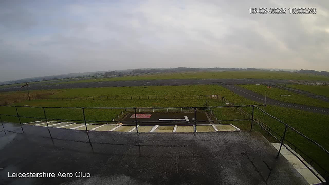 A view from a webcam positioned on a balcony overlooking an airstrip. The scene is mostly grassy with a few patches of dark asphalt. Several sections of the runway are visible, along with a fenced area beneath the balcony. The sky is overcast with gray clouds, and the atmosphere appears calm.