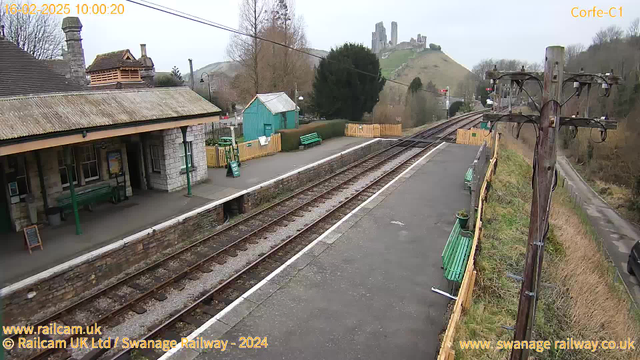 A view of a train station platform with stone and wooden structures. The platform features green benches and a sign. In the background, there is a hill with ancient ruins visible at the top. The sky is overcast, and there are trees surrounding the area. The railway tracks are visible, running parallel to the platform.