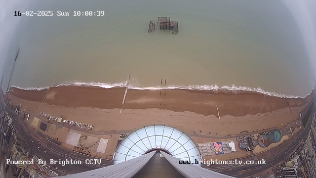 Aerial view of a beach with brown sand and gentle waves lapping the shore. A dilapidated pier structure is visible in the water, and there are several small objects or structures on the beach. The image is taken from a high vantage point, showing part of a building or observation platform with a curved glass roof at the lower center. The sky appears overcast. In the background, a road runs parallel to the beach with various colored structures, possibly amusement park rides or attractions, visible along the shoreline. The date and time are displayed in the top left corner.