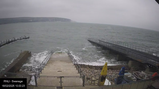 A cloudy scene of a coastline with choppy grey waters. In the foreground, a concrete ramp leads down to the water, bordered by metal railings. Adjacent to the ramp, there are kayaks in bright colors (yellow, blue, and red) stored on the pebbled shore. To the left, a jetty extends into the water with several posts, while the coastline in the background features cliffs. The image appears to be taken during the day with overcast skies.