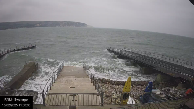 A cloudy scene at a seaside location features choppy waves lapping at the shore. In the foreground, a concrete ramp leads down to the water, outlined by a metal railing. To the right, a small dock extends over the water, with waves crashing against it. Several boats, including a yellow one, are lined up along the pebbled shore. In the background, green hills gently rise, while a rocky cliff is visible in the distance. The mood is overcast and windy.