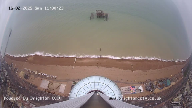 A view from a high vantage point overlooking a sandy beach, with gentle waves lapping at the shore. In the distance, a broken pier extends into the water. The beach is lined with various structures, including some that appear to be amusement rides or facilities. A clear sky is visible above the scene. The current date and time are displayed in the upper left corner, indicating it is February 16, 2025, at 11:00 AM. The bottom of the image shows a curved glass structure, likely part of an observation deck.