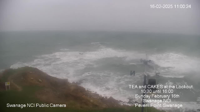 A view of the ocean on a cloudy day, with waves crashing against a rocky shoreline. The sky is grey, indicating overcast weather. In the foreground, there is a sign partially visible on the left, near the edge of a grassy area. In the distance, the water appears turbulent with white frothy waves. A weather vane is positioned to the right, pointing north. The date and time are displayed in the top right corner, along with text promoting an event for tea and cakes at the lookout.