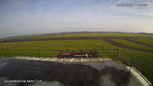 A view from a webcam at Leicestershire Aero Club showing a long grassy field adjacent to a runway. The runway is marked with white lines and surrounded by a safety fence. In the distance, there are more green fields and hills under a partially cloudy sky. The image features flat surfaces and a railing in the foreground, with a glimpse of wet pavement. The time and date display in the corner indicates it is 11:00 AM on February 16, 2025.