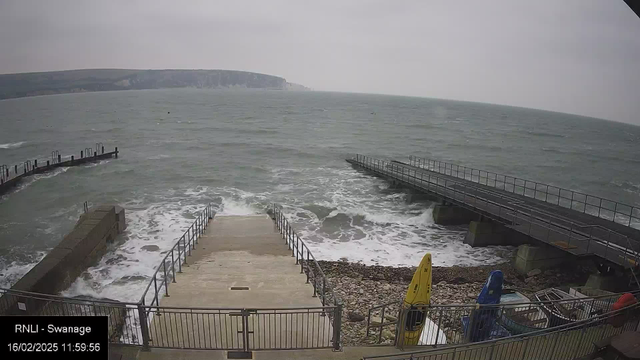 A view of a coastline with choppy gray waters and overcast skies. In the foreground, there are steps leading down to the water, flanked by a railing. To the left, there is a wooden pier jutting out into the sea, while a second, shorter pier is visible to the right. At the bottom right, there are several kayaks; one is yellow, and another is blue. The rocky shoreline is partially covered by water, and distant cliffs can be seen in the background.