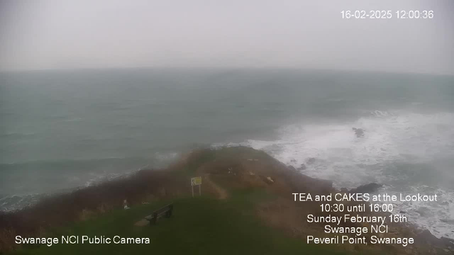 A coastal view is depicted with turbulent, grayish-green waters crashing against the rocky shoreline. In the foreground, there is a grassy area with a park bench, and a nearby sign indicating an event. The sky is overcast, leading to a gloomy atmosphere with limited visibility. The watermark in the bottom left corner reads "Swanage NCI Public Camera," while text on the right announces a tea and cakes event at Peveril Point, Swanage, on February 16th, from 10:30 to 16:00.