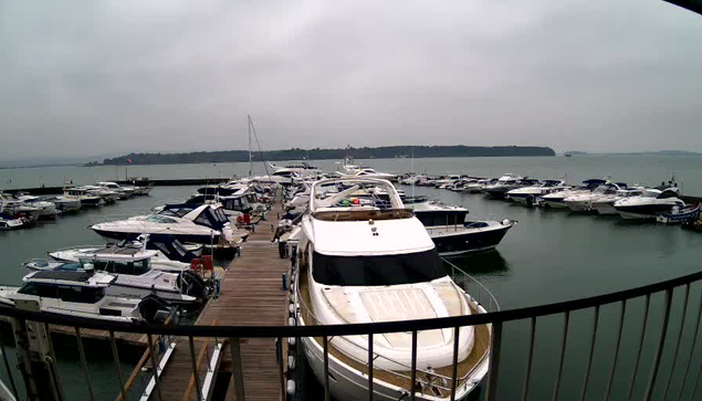 A view of a marina on a cloudy day, with numerous boats docked along a wooden pier. In the foreground, a large white yacht is prominent, with several smaller boats visible around it. The water appears calm, and there are hills in the background shrouded in mist. The overall atmosphere is quiet and serene, with a muted color palette due to the overcast skies.