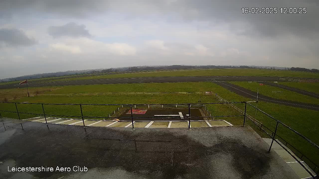 A cloudy day at an airfield viewed from a webcam. In the foreground, there is a railing along a walkway. Below the walkway, a grassy area is visible, with a few patches of brown indicating the ground. Ahead, a long runway stretches across the image, bordered by a wooden fence on the far side. The sky is overcast, creating a muted lighting effect, and the date and time are displayed in the corner.