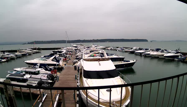 A view of a marina on a cloudy day, featuring numerous boats moored at the docks. The foreground shows a wooden walkway leading to the water, surrounded by various yachts and smaller vessels. The water is calm with a grayish hue, and a distant shoreline is visible in the background under a cloudy sky.