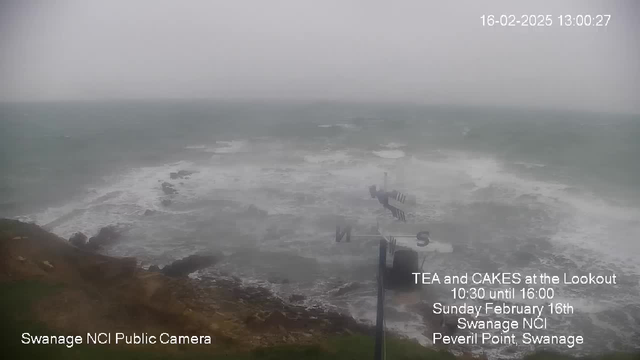 A cloudy, overcast scene showing turbulent ocean waves crashing against rugged rocks along a coastline. The sky is gray, suggesting a stormy atmosphere, and visibility is low. In the foreground, a weather vane with directional indicators stands on a post. Text overlay indicates a public event for tea and cakes happening at a lookout point in Swanage. The image appears to be captured from a public webcam.