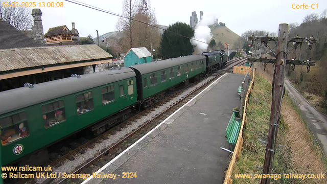 The image shows a train station in Corfe, England, with a green steam train departing. The train has several green carriages, with people visible through the windows. In the background, there is a hill with ruins visible. The station features a sloped roof building on the left and several green benches along the platform. There are trees and a wooden telephone pole on the right. The atmosphere appears overcast, suggesting a cool day.