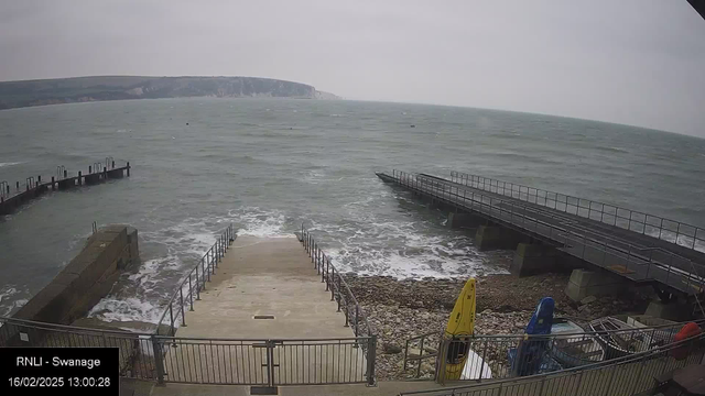 A view of a seaside location on a cloudy day. In the foreground, there are concrete steps leading down to the water's edge, bordered by a metal railing. To the right, two colorful kayaks, one yellow and one blue, are stored against a railing. In the background, the choppy sea extends to the horizon, where a distant cliff is visible. The overall atmosphere suggests a windy day at the coast.