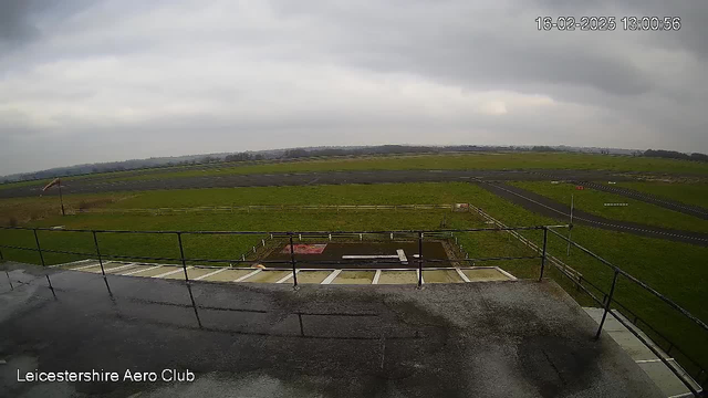 A view from an elevated position at the Leicestershire Aero Club, showing a vast expanse of green grass under a cloudy sky. In the foreground, there is a flat rooftop area with railing. Below, there is an airfield with a runway marked by black asphalt and white lines. A few scattered fences are visible in the distance, and a flag is gently blowing in the wind on the left side of the image. The scene has a tranquil, overcast atmosphere.