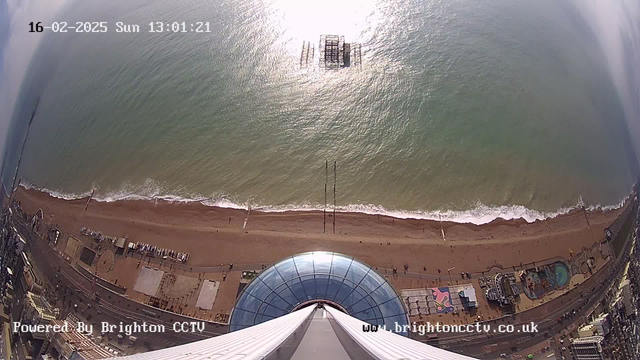 An aerial view from a tall structure overlooking a beach with golden sand and gentle waves. The sunlight reflects on the water, creating a shimmering effect. In the distance, there is a partially submerged pier. The beach is dotted with people and features colorful amusement rides and structures along the shore. The scene captures a vibrant seaside atmosphere on a sunny day. The image is timestamped, indicating the date and time of the view.