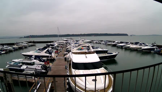 A marina scene featuring numerous yachts and boats docked at a wooden pier. The sky is overcast with a gray hue. In the background, there is a calm body of water, and on the far side, green hills are visible. The boats vary in size and style, with some sporting colorful covers and equipment on board. The scene conveys a tranquil and slightly cloudy day at the marina.