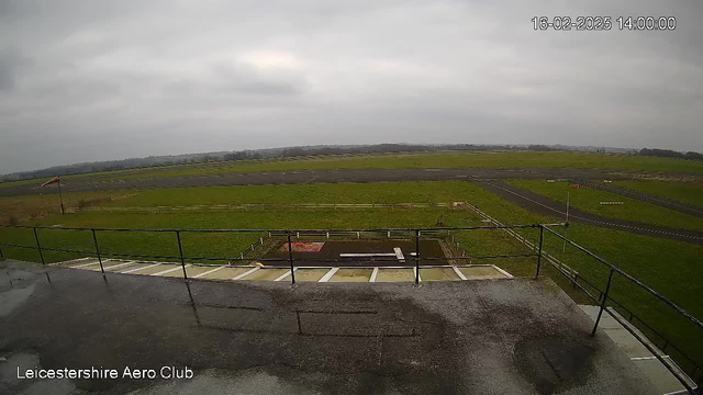 A cloudy sky fills the background over a large open field. In the foreground, there is a balcony railing surrounding a flat rooftop, with water pooling on the surface. Below, the green grass and a dirt area are visible, separated by a wooden fence. A section of the ground appears to be marked for runway use, with visible markings and small signs. In the distance, the horizon is somewhat obscured by the gray sky.