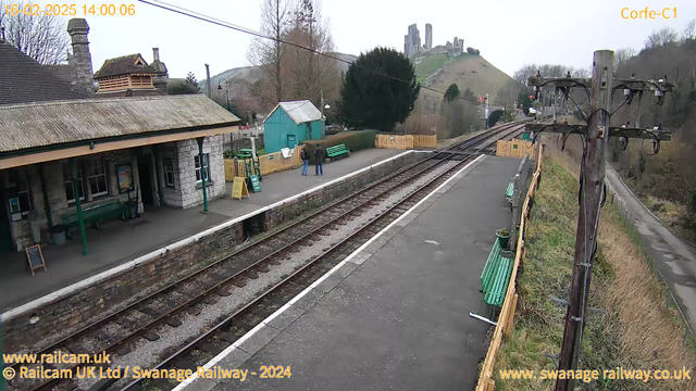 A train station platform with a stone building on the left, featuring a sloped roof and several windows. Two people stand on the platform, facing towards the left side of the image. There are green benches along the platform, and a small turquoise building can be seen in the background. To the right, there are railway tracks leading away from the camera, bordered by gravel. In the distance, a hill rises, topped by ruins of a castle. A wooden utility pole with wires stands near the platform's edge, and a narrow road runs alongside the hill in the background. The sky is overcast.