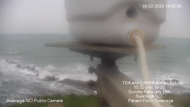 A blurred image showing a coastline with waves crashing against rocks. In the foreground, there is a white object, possibly a container, positioned on a gray platform. The weather appears overcast and foggy, creating a muted atmosphere with limited visibility. Text on the image indicates the date and time, along with an event advertised at "The Lookout" in Swanage.