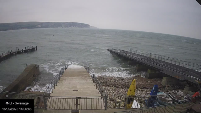 A coastal scene showing a view of the sea with a rocky shoreline. In the foreground, a set of concrete steps leads down to the water, surrounded by a metal railing. To the left, there is a wooden pier extending into the sea. On the right, there are several colorful kayaks—yellow, blue, and red—secured on the ground. The sea is choppy with white waves, and the sky is overcast, giving a muted appearance to the landscape. A cliff can be seen in the distance against the horizon.