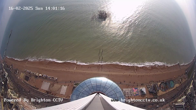 Aerial view of a sandy beach with gentle waves lapping at the shore. The image shows a broad stretch of beige sand with small figures of people walking along the water's edge. On the left side, there is a group of wooden poles standing in the water, leading to a partially submerged pier structure. Below the beach is a circular, glass-roofed building, contrasting with the surrounding sandy area. The scene is illuminated by sunlight reflecting off the water, creating a shimmering effect.