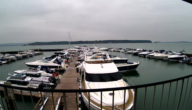 A marina scene on a cloudy day, featuring numerous boats docked at a pier. Several motorboats and yachts of varying sizes are lined up in the water, reflecting the overcast sky. The background shows a sliver of green hills against the gray horizon, and the water appears calm with gentle ripples. A wooden walkway extends across the foreground, leading to the boats.