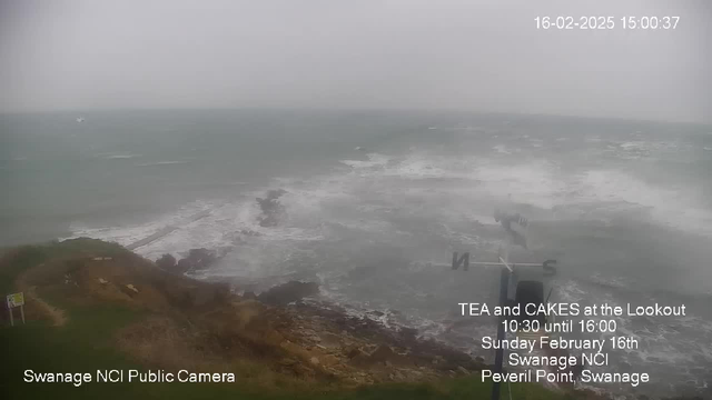 A cloudy seaside view shows turbulent waves crashing against a rocky coastline. In the distance, a small boat is visible on the water. The scene appears hazy due to fog or mist, obscuring visibility further out to sea. At the bottom of the image, there's a sign indicating a tea and cake event at a lookout point on February 16th, along with markers identifying the camera's location at Peveril Point, Swanage.