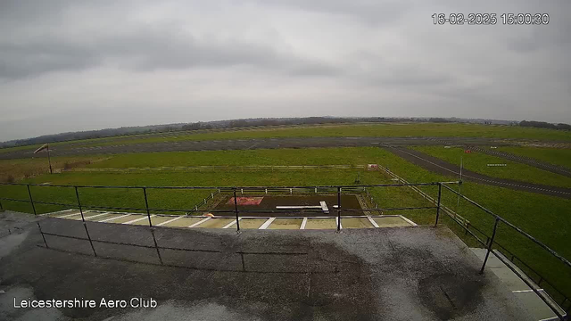 A view from a webcam at Leicestershire Aero Club, showing a wide expanse of grassy airfield under a cloudy sky. In the foreground, there is a wooden railing, and a small section of the ground with white stripes visible. The airstrip, which appears grey and paved, extends into the distance. Signs and a fence are visible along the edge of the field, with slightly rolling terrain beyond. The image is timestamped February 16, 2025, at 15:00.