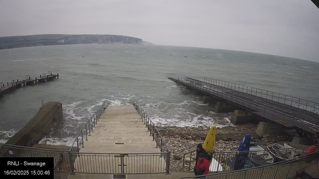 A coastal scene shows a stairway leading down to the shoreline, with waves gently crashing at the base. Two individuals are walking down the steps, while another person is ascending. To the left, there is a solid structure on the water's edge, with a railing along the stairway. In the background, a pier extends into the sea, and rugged cliffs can be seen in the distance under a grey sky. Several boats and a yellow kayak are visible at the shore.