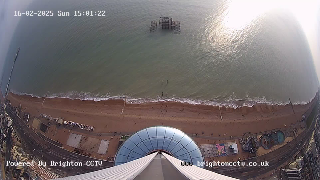 A high-angle view looking down from a tall structure over a sandy beach and the sea. The beach is mostly empty with a few people visible along the shoreline. In the water, a partially submerged pier structure is seen. The coastline features a road with structures, including a circular amusement area and a swimming pool. The scene is bathed in soft sunlight reflecting off the water, and the sky is partly cloudy. The date and time are displayed in the top left corner of the image.