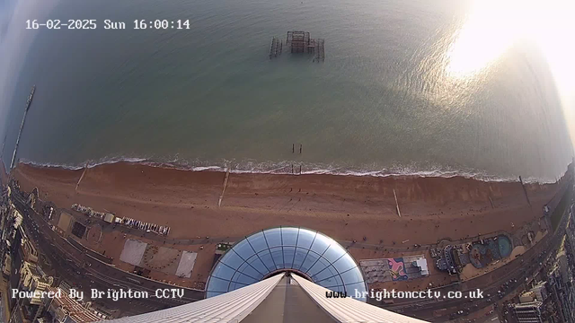 A high-angle view of a sandy beach extending into the sea, with gentle waves lapping at the shore. In the foreground, a large circular structure, possibly a viewing platform, is visible, reflecting sunlight. To the left, a long wooden pier stretches into the water, while remnants of an old pier are seen further out. The shoreline is dotted with colorful beach huts, and a small amusement area is visible, with people scattered across the beach. The date and time display is positioned at the top left of the image.