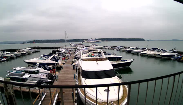 A marina filled with various boats is visible, with many larger yachts docked in a calm body of water. The sky is overcast, giving a gray tone to the scene. A wooden deck extends along the edge of the marina, with the foreground featuring a larger white boat, displaying a covered cockpit area. The boats are lined up in neat rows, some with colorful coverings. In the background, the shoreline is faintly visible, along with a few distant boats further out on the water.