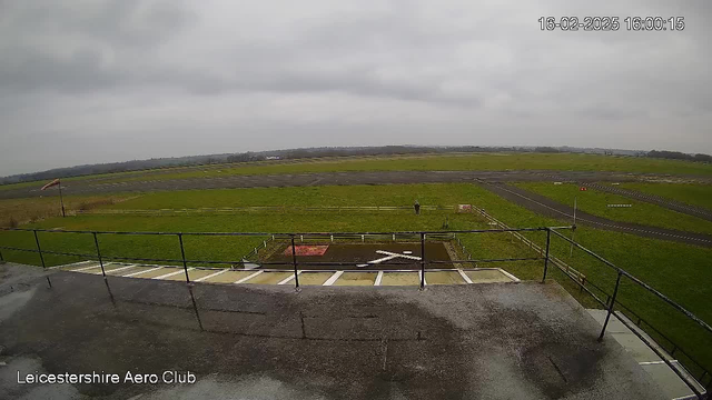 A view from a webcam overlooking an airstrip. The scene is mostly cloudy with a gray sky. In the foreground, there is a fenced area and a person standing at a distance. An X-marked area is visible on the ground, likely indicating a landing or takeoff spot. The landscape features green grass surrounding the airstrip, and a small wind indicator can be seen on the left side of the image. The airstrip itself is dark and paved, leading into the distance.