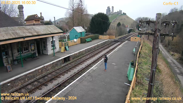 A view of Corfe Castle station, featuring a stone building with a sloped roof on the left, and a green wooden shed in the background. A person stands on the platform, looking at a device. Two railway tracks run through the scene, with a few benches along the platform. In the distance, the ruins of Corfe Castle can be seen atop a hill, surrounded by trees and grassy slopes. The sky is overcast, indicating a cloudy day.