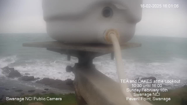 A webcam view shows a blurred image of a white cylindrical object, possibly a container, positioned on a ledge. Below, rough ocean waves crash against rocks, indicating a coastal scene. The sky appears overcast, and the overall image has a foggy quality. Text in the corner provides details about a tea and cakes event at a lookout point in Swanage.