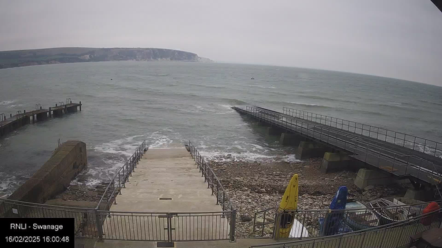 A waterfront scene showing a view of the sea. There is a set of stairs leading down to the water, flanked by rocky shore and a metal railing. To the left, a wooden jetty extends into the ocean, and to the right, a second jetty is visible. Several colorful kayaks in yellow, blue, and red are positioned on the ground near the water’s edge. The sky is overcast, and the water is choppy with small waves. Cliffs can be seen in the background. 