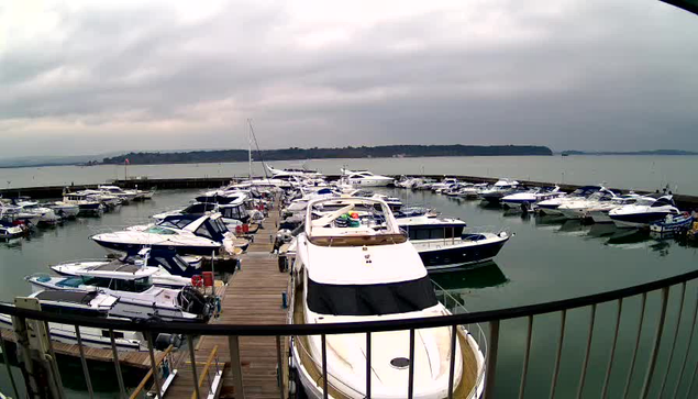 A marina with numerous boats docked in a tranquil body of water. The scene is under overcast skies, with grey clouds overhead. The boats vary in size, and some are tied to wooden docks. In the distance, a shoreline can be seen with trees and hills. The water reflects the muted colors of the sky. A metal railing is visible in the foreground, framing the view of the marina.