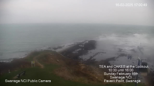 A cloudy coastal scene featuring rocky areas along the shoreline. The sea is calm with gentle waves lapping against the rocks. In the foreground, there is a grassy area with a bench on the left side, and a signpost. The image captures a muted color palette, primarily grays and blues, typical of an overcast day. Text at the bottom indicates "TEA and CAKES at the Lookout" with details about the event and its location at Peveril Point, Swanage.